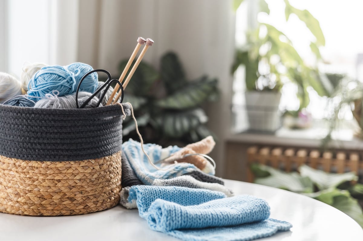 Canasta con lanas, agujas y piezas tejidas sobre mesa con plantas