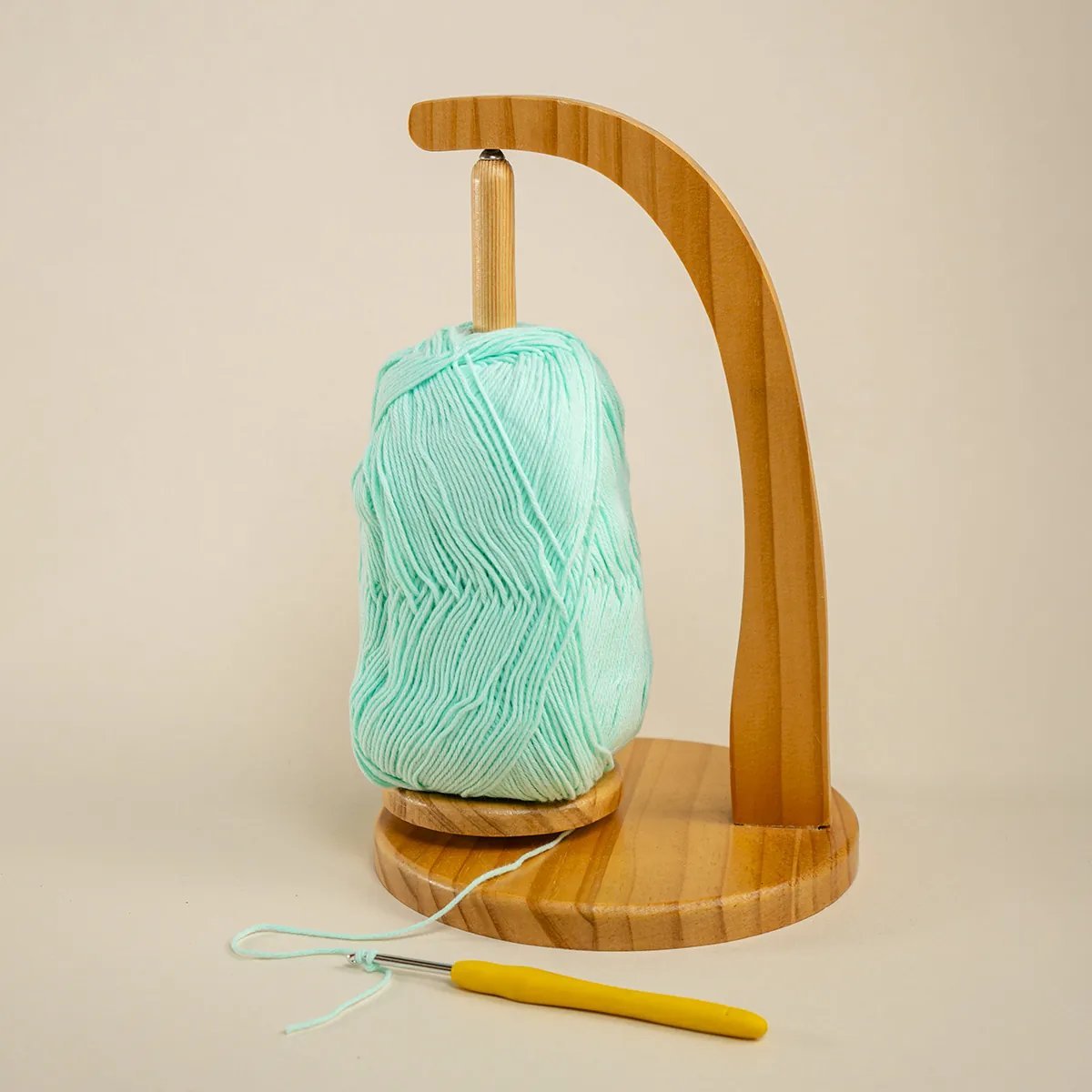 Soporte de madera en forma de arco para ovillos con un ovillo de lana color menta y un crochet amarillo, fondo beige
