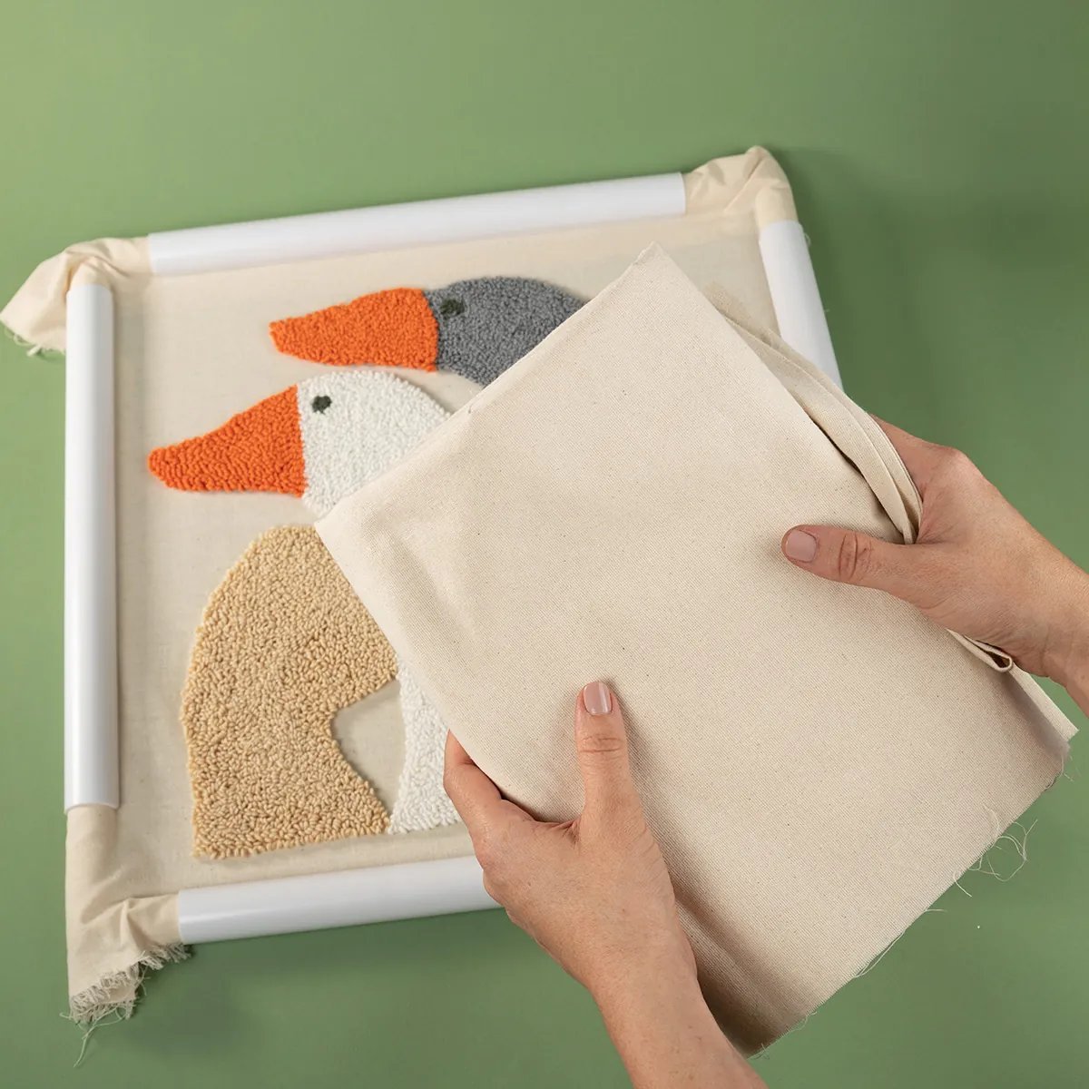Manos sosteniendo un trozo de tela beige sobre un cuadro punch needle con un pez bordado en colores sobre fondo verde