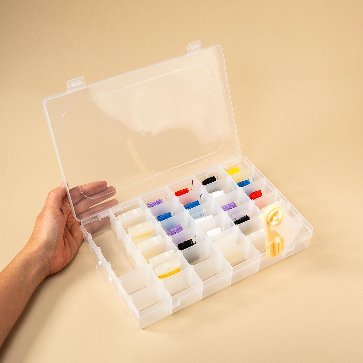 Mano sosteniendo una caja organizadora de plástico transparente abierta con compartimentos llenos de carretes de hilo de colores para costura y manualidades